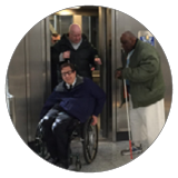Three men exit an elevator, one using a wheelchair and one with a cane.